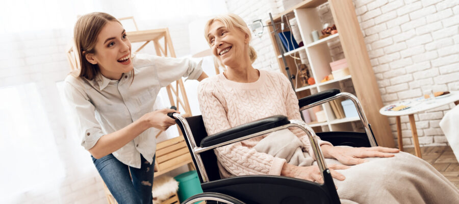 Girl is nursing elderly woman at home. Girl is riding woman in wheelchair. They are happy.